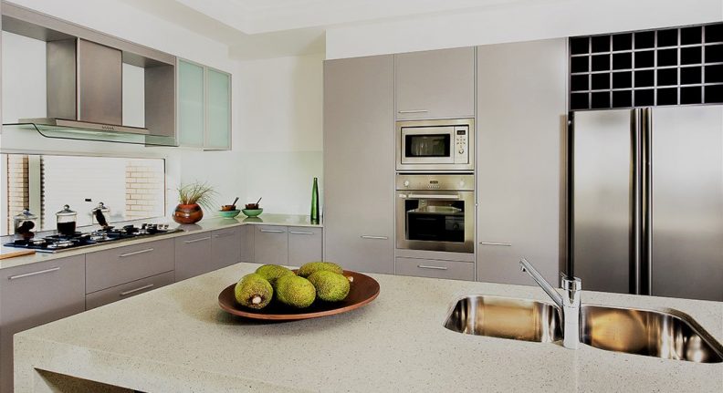 Counter top made in ceramic seen closely with fruits on a brown color clay dish next to the modern  silver faucet attached to the sink, the silver refrigerator and the wall oven with pantry cupboards next to the gas stove